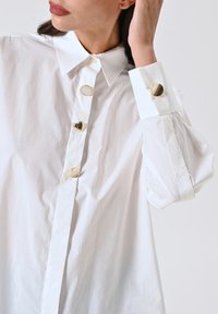 Camicia bianca con colletto classico. Presenta bottoni dorati, inclusi quelli a forma di cuore, e maniche a sbuffo con polsini. Tessuto dalla consistenza liscia.
