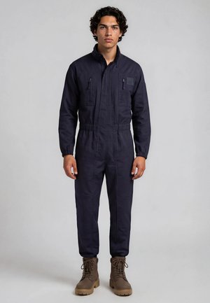 Homme aux cheveux bouclés foncés portant une combinaison bleu marine avec des poches zippées sur la poitrine et des bottes marron à lacets, debout devant un fond uni.