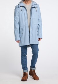 Vaaleansininen vedenpitävä takki, jossa on harmaa huppu, kaksi etutaskua, vetoketjullinen rintatasku ja nepparikiinnitys. Päällä harmaa paita ja farkut.