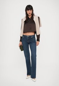 Camisa de botones corta color marrón claro, vaqueros acampanados azul oscuro y zapatos crema de punta puntiaguda. Suéter beige grande colgado sobre los hombros.