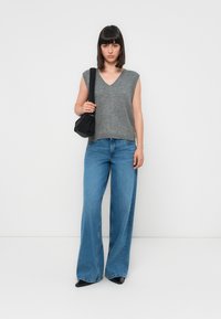 Grijze mouwloze gebreide top met een V-hals, gecombineerd met brede blauwe jeans. Model houdt een kleine zwarte handtas vast en staat tegen een witte achtergrond.