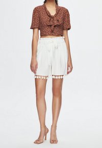 Cropped brown floral blouse with a bow detail and short sleeves, paired with white shorts featuring a tie waist and decorative pom-pom trim.