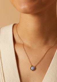 Gouden ketting met een ronde blauwe stenen hanger, geplaatst in een gestructureerd gouden frame, rustend op een lichte huidtint.