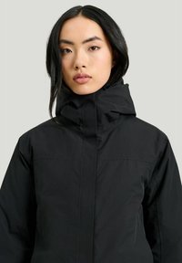 Svart isolerad jacka med hög krage och dold dragkedja. Materialet verkar texturerat, designat för värme och väderbeständighet.