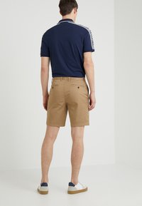 Polo shirt marine avec des accents blancs, associé à des shorts beiges. Les chaussures sont blanches avec des détails navy et gris. Vue arrière mettant en évidence l'ajustement et la longueur.