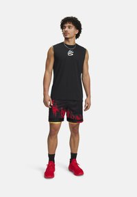 Top senza maniche nero con logo, abbinato a pantaloni corti neri con stampa a fiamma rossa e accenti gialli. Indossato con scarpe da ginnastica rosse e calzini neri.