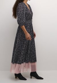 Maxi šaty v šedé barvě s černým leopardím vzorem, s nabíraným pasem, plisovaným růžovým tylovým lemem a tříčtvrtečními balónovými rukávy. Černé kotníkové boty.