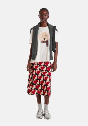 Camiseta de manga corta color crema con un gráfico de perro, suéter sin mangas gris y falda hasta la rodilla con un patrón de corazones rojos y crema. Zapatillas grises y calcetines.