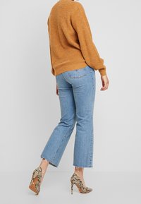 En person i en brun tröja, ljusblå cropped jeans och högklackade skor i ormskinn stående framför en helt vit bakgrund.