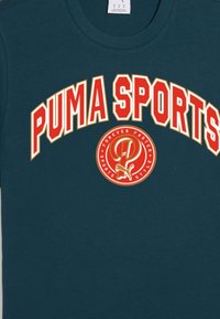 Jūrinis marškinėliai iš medvilnės mišinio su užrašu "PUMA SPORTS" ryškiu raudonu tekstu ir apskrito logotipo grafika centre su pabrėžtu tekstu.
