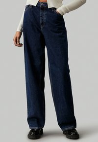 Jeans de mezclilla de color azul oscuro y pernera ancha con una cintura alta, que cuentan con bolsillos frontales y costuras de contraste. Frontal plano y ajuste holgado.