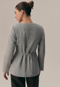 Femeie cu părul prins în coc, purtând o jachetă gri cu model în pepită, cu detaliu de curea în spate și cercei tip cerc, cu fața întoarsă cu spatele către cameră.