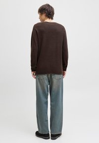 Personen står med ryggen til, iført en mørkebrun sweater, løse blå jeans, sorte sko og en tatovering på bagsiden af nakken.