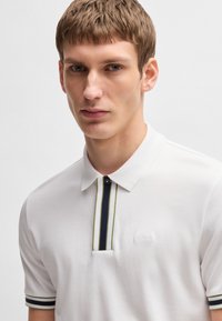 Biała koszulka polo wykonana z bawełny, z klasycznym kołnierzykiem, prążkowanymi akcentami w kolorze granatowym i zielonym oraz małym wytłoczonym logo na piersi.