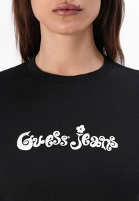 Sweatshirt preto com o logótipo "Guess Jeans" em branco, com decote arredondado e detalhes em malha. Material de algodão suave.