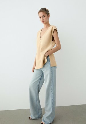 Giovane donna che indossa un top color sabbia senza maniche con scollo a V e jeans chiari a gamba larga, in piedi con una mano in tasca su sfondo semplice.