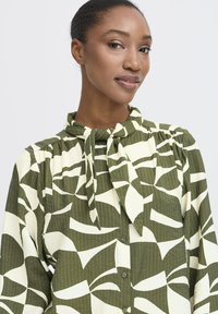 Kvinde med mørkt hår samlet bagpå, iført en grøn og hvid mønstret bluse med en bundet hals_sløjfe, som ser selvsikkert på kameraet.