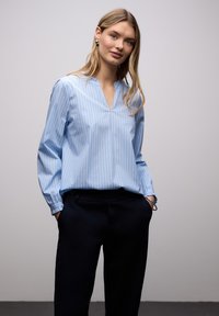 Blouse bleu clair à rayures blanches verticales, sans col, manches longues et coupe décontractée, associée à un pantalon bleu marine foncé.