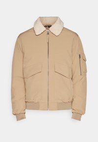 Chaqueta bomber marrón claro con cuello de borrego, que presenta un cierre frontal, dos grandes bolsillos con solapa y un bolsillo en la manga con cremallera.