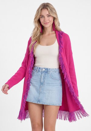Ung kvinde iført en lyserød cardigan med frynser, hvid tanktop, lys denim nederdel og lange lyserøde frynøre, smilende med løst blond hår.