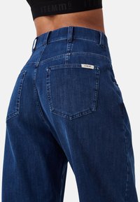 Jeans in denim blu scuro con vita alta, due tasche posteriori e un'etichetta bianca con "ITEM m6" sulla cintura.