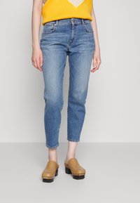 Modré klasické džíny s mírným zúžením, které jsou nošeny s žlutým pleteným topem s výstřihem do V. Béžové dřeváky mají masivní černou podrážku a detail prošívání.