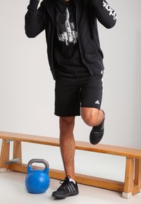 Muž v černém sportovním oblečení balancuje na jedné noze na dřevěné lavičce, vedle něj leží modrý kettlebell.