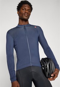 Homme portant un maillot de cyclisme à manches longues bleu marine et un short rembourré noir, tenant un casque de vélo noir devant un fond uni.