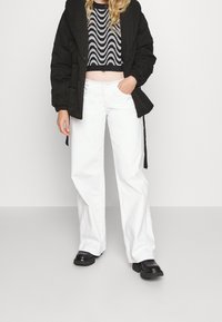 Svart puffjacka med huva, en kort svart och silver vågmönstrad tröja, vita vida byxor och kraftiga svarta stövlar.