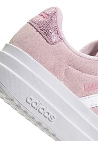 Zapatilla de gamuza rosa con un talón metálico rosado texturizado, entresuela blanca y tres rayas blancas. Presenta un logotipo de la marca en relieve en el costado.