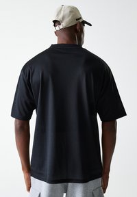 T-shirt en mesh noir à manches courtes, avec une coupe décontractée et une texture perforée subtile. Vue de dos, porté avec une casquette beige clair.