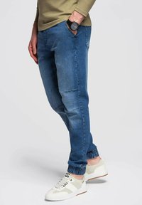 Joggers de mezclilla azul con puños elásticos, que presentan una textura suave. Combinados con zapatillas blancas y gris claro y un reloj negro.