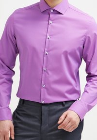 Fioletowa, dopasowana, koszula na długi rękaw z guzikami w kontrastującym kolorze oraz gładką, teksturowaną tkaniną. Zestawiona z ciemnymi spodniami.