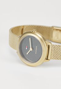 Montre dorée avec un cadran sombre scintillant, présentant des index d'heure minimalistes et un bracelet en maille dorée. Logo affiché sous la marque des 12 heures.