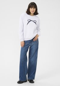Lichtgrijze sweatshirt met een marineblauwe strikgrafiek op de borst, gecombineerd met wijde blauwe denim jeans. Zwarte schoenen maken de outfit compleet.