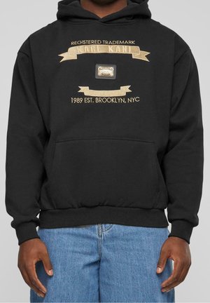 Czarna bluza z kapturem z złotym haftem z napisem "Registered Trademark Karl Kani 1989 Est. Brooklyn, NYC", noszona z niebieskimi dżinsami.