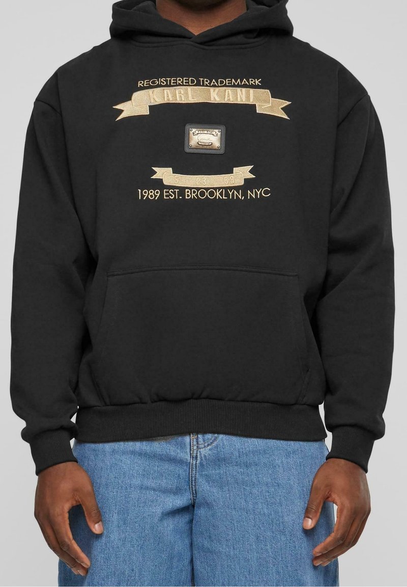 Sweat à capuche noir avec broderie dorée indiquant « Registered Trademark Karl Kani 1989 Est. Brooklyn, NYC », porté avec un jean bleu.