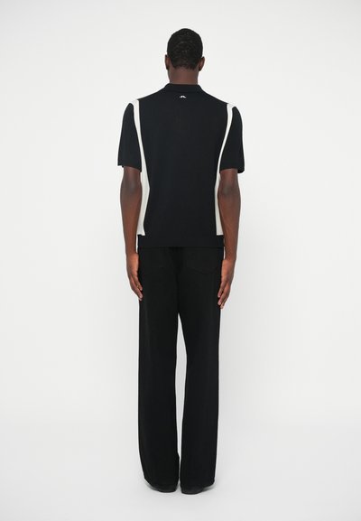 Tricou polo negru cu accente albe pe laterale, mâneci scurte și guler cu nervuri. E combinat cu pantaloni negri largi, creând o siluetă elegantă.