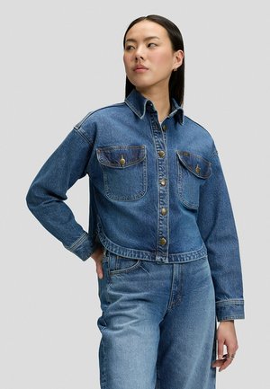 Femme portant une veste en jean bleue avec des poches avant et un jean taille haute assorti, debout, une main sur la hanche, regardant sur le côté.