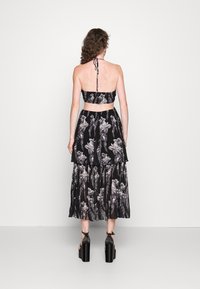 Nainen, jolla on kihara hiuksia, pukeutuneena mustiin korkokenkiin, mustaan kukalliseen olkaimettomaan crop-toppiin sekä yhteensopivaan kerrokselliseen midi-hameeseen, kääntynyt pois.