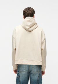 Άτομο που φοράει μια ανοιχτό μπεζ φούτερ με κουκούλα καλυμμένη και μπλε τζιν παντελόνι καρό, με θέα από πίσω πάνω σε μια απλή φόντο.