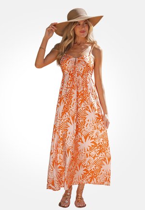 Frau mit breitkrempigem Strohhut und ärmellosem orange-weißem Blumenmaxikleid, hält mit einer Hand den Hutrand, steht barfuß.