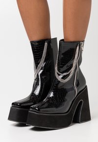 Černé lakované kotníkové boty s krokodýlí texturou, stříbrnými řetízkovými detaily, dvojitými zipy a silnou černou platformou.