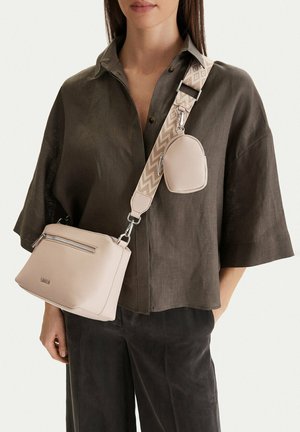 Kobieta ubrana w ciemną, luźną koszulę i spodnie, niosąca beżową torbę na pasku przez ramię z szerokim, wzorzystym paskiem oraz małą przypiętą saszetką.