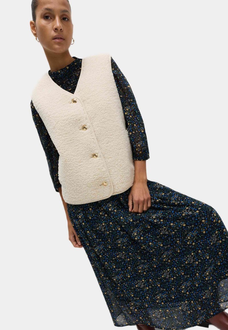 Chaleco texturizado crema con botones dorados, usado sobre un vestido largo con estampado floral oscuro y acentos en azul y amarillo.
