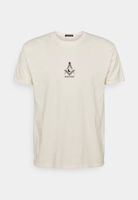 Bílý krátký rukáv T-shirt s malým černým geometrickým vzorem a textem "KAOTIKO" uprostřed na hrudi.