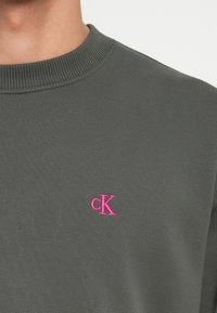 Tmavosivá mikina s rebrovaným okrúhlym výstrihom. Na hornej ľavej strane hrude sa nachádza malé ružové vyšívané logo "ck".
