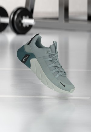 Zapatilla deportiva Nike de entrenamiento en tonos teal claro y gris con suela gruesa, exhibida flotando sobre el suelo del gimnasio con mancuernas desenfocadas en el fondo.