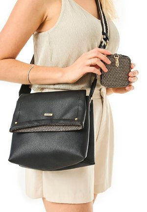 Donna in abito beige che tiene una piccola pochette intrecciata con cerniera e indossa una borsa a tracolla in pelle nera con pattina decorata.