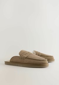 Pár hnedých semišových šliapacích papúč s mäkkým bielym vnútorným podšívkou na jednofarebnom svetlom pozadí.